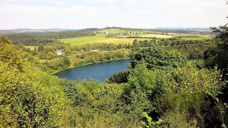 A picturesque view of a tranquil lake, surrounded by green trees and gentle hills. The sky is clear and the landscape appears peaceful.