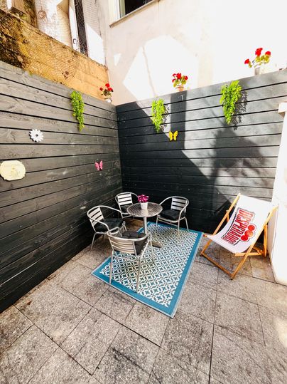A cozy courtyard with gray walls and a blue carpet. There is a round table with chairs and some flowers on the wall.