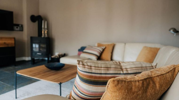 Ein gemütliches Wohnzimmer mit einem beigen Sofa und bunten Kissen. Ein niedriger Tisch und ein Holzofen runden die entspannte Atmosphäre ab.