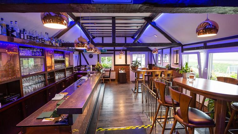 A modern bar with wooden furniture and stylish lighting elements. In the background, windows with a view of the outdoors and a selection of beverages are visible.