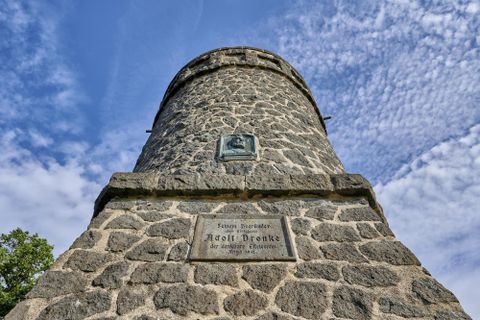 Ein historischer Steinturm mit einer Gedenktafel an der Front. Der Himmel ist blau mit einigen Wolken.