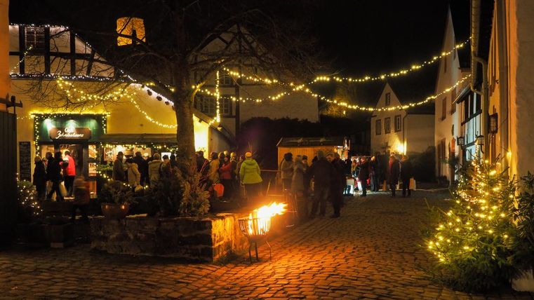 Ein festlich beleuchteter Platz mit Menschen, die sich versammeln. Weihnachtslichter hängen über dem Kopf und ein geschmückter Baum steht in der Ecke.