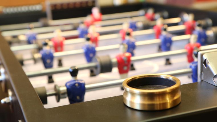 Ein Tischfußballspiel mit Figuren in Rot und Blau. Darauf liegt ein runder, goldfarbener Becher.