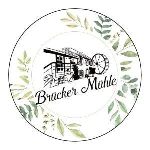 Ein runder Teller mit dem Schriftzug "Brücker Mühle" und einer stilisierten Abbildung einer Mühle. Umrahmt von grünen Blättern wirkt das Design naturverbunden und einladend.