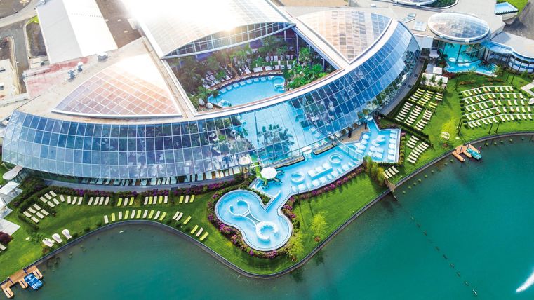 Een modern hotel met een glazen façade en een groot zwembadgebied. Omringd door water en ligbedden, biedt het een uitnodigende plek om te ontspannen.