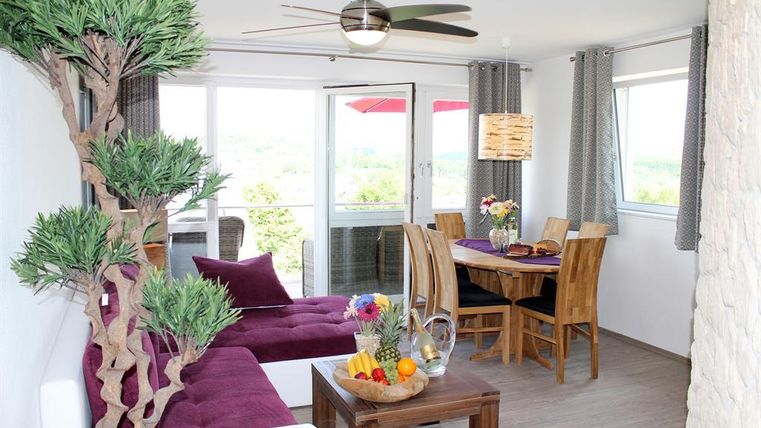 Ein modernes Wohnzimmer mit einem roten Sofa, einem Tisch mit frischen Früchten und einer Esstischgruppe. Große Fenster lassen viel Licht herein und bieten einen schönen Ausblick.