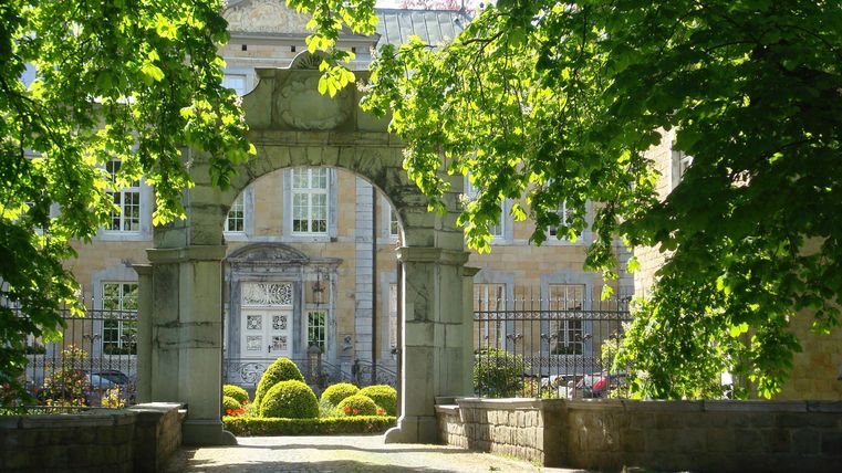 Ein schöner Eingang mit einem Bogen aus Stein und umgeben von Bäumen. Im Hintergrund sieht man ein Gebäude mit Fenstern und einem gepflegten Garten.