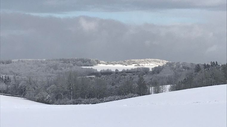 A snow-covered landscape with gentle hills and snow-laden trees. The sky is cloudy and conveys a serene atmosphere.