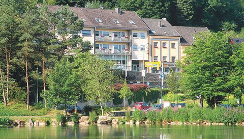 Een rustige vijver met aangrenzende bomen en grasvelden. Op de achtergrond zijn meerdere gebouwen te zien die door de natuur omringd zijn.
