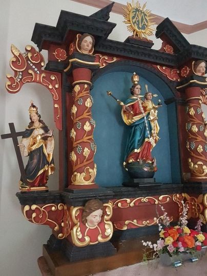 Ein kunstvolles Altarbild mit farbenfrohen Skulpturen von Heiligen. Es zeigt eine Statue der Jungfrau Maria mit dem Kind und Blumenarrangements davor.