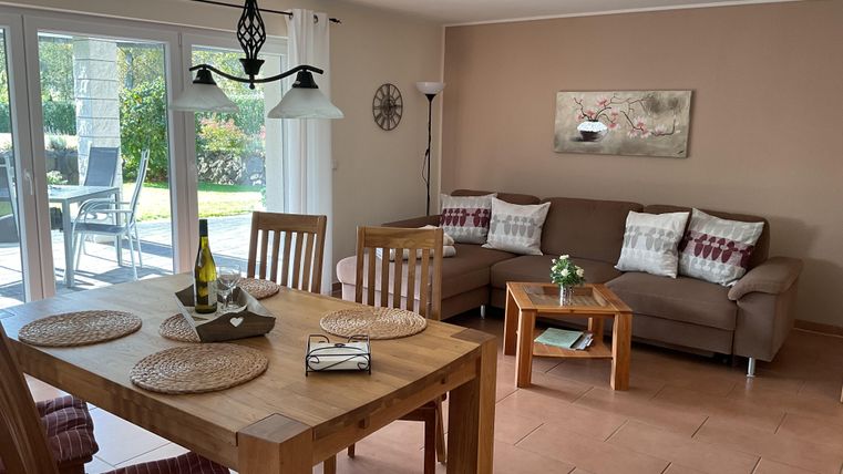 A cozy living and dining room with a wooden tray and a bottle of wine on the table. Large windows let in plenty of daylight and offer a view of the garden.