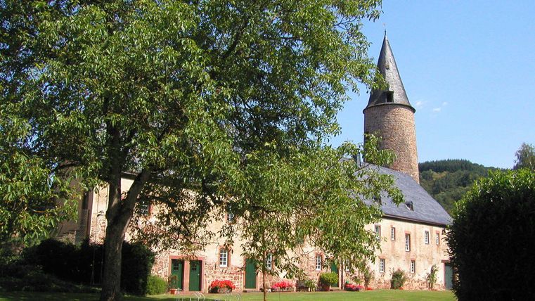 Een historisch gebouw met een spits toren en een grote, groene weide. Op de voorgrond staan enkele tafels en stoelen onder een boom.