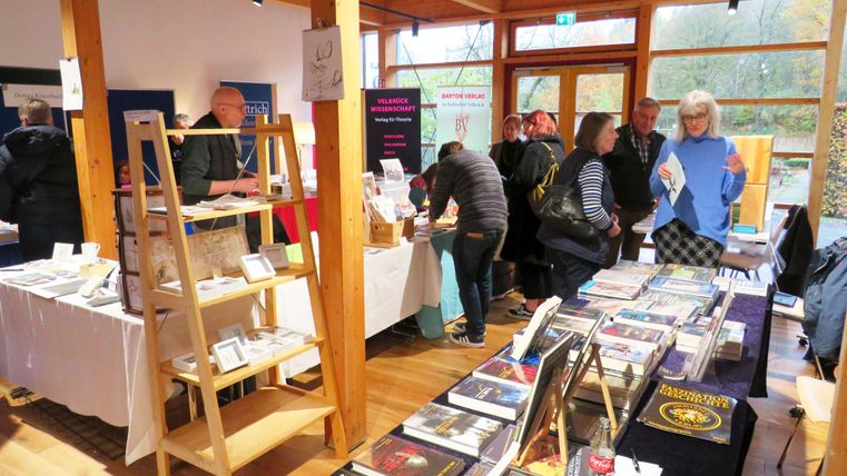 Een drukke markt met verschillende kraampjes en tafels. Mensen praten met elkaar en bekijken een verscheidenheid aan boeken en kunstwerken.