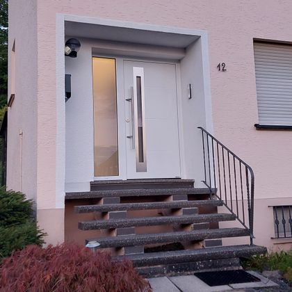 Eine moderne Haustür in einer hellen Fassade. Die Eingangsstufen sind von einem kleinen Garten umgeben.