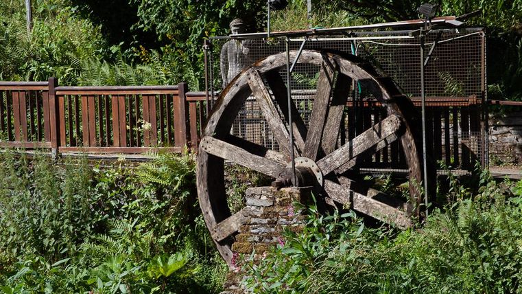 Ein altes Wasserrad steht inmitten von üppigem Grün. Im Hintergrund ist eine Holzkonstruktion zu sehen.