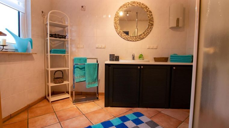 A modern bathroom with a large mirror and elegant blue towels. The room is bright and tidy with a stylish shelf and tiled floor.