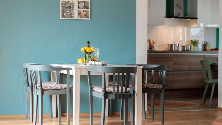 Ein stilvoller Esstisch mit Stühlen, umgeben von einer blauen Wand. Auf dem Tisch stehen eine Blumenvase und mehrere Getränke.