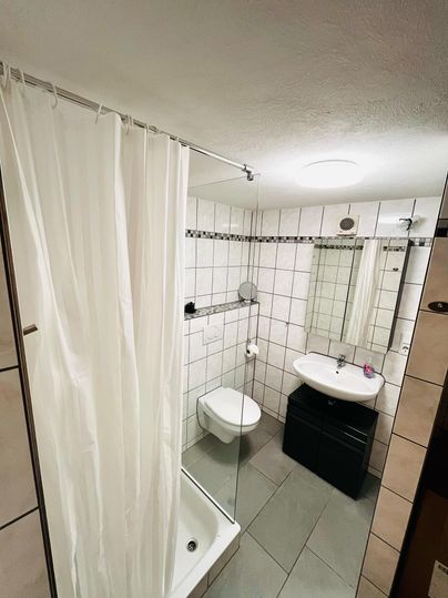 A modern bathroom with a shower, a toilet, and a sink. The walls are decorated with white tiles and a large mirror.