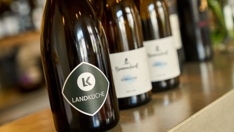 Eine Flasche Wein mit dem Etikett "LANDKÜCHE" steht im Vordergrund. Im Hintergrund sind weitere Weinflaschen mit weißen Etiketten zu sehen.