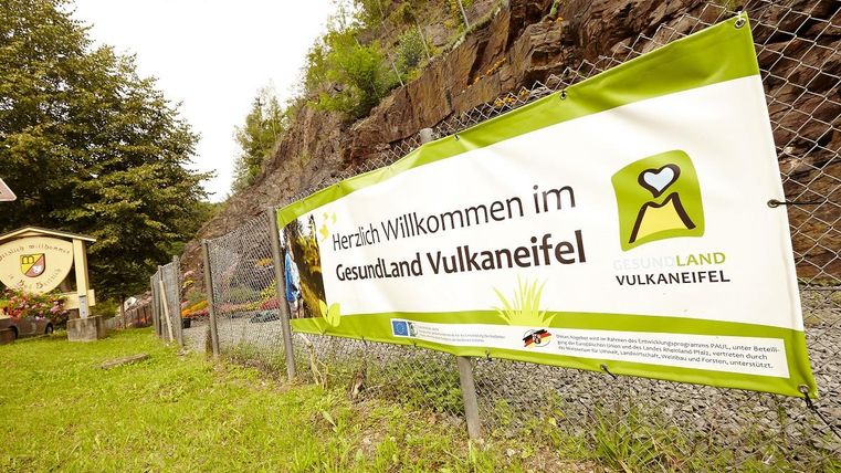 Ein Willkommensschild für das Gesundheitsland Vulkaneifel steht an einer Zaun. Im Hintergrund sind Bäume und Felsen zu sehen.