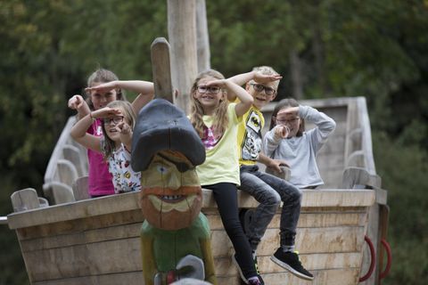 A group of children is posing on a play ship, most of them are saluting with their hands. In the foreground, there is a large wooden figure of a pirate.