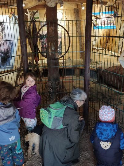 Eine Gruppe von Kindern und einer älteren Frau steht vor einem Käfig in einem Tiergehege. Im Hintergrund sind Bilder an der Wand sichtbar.