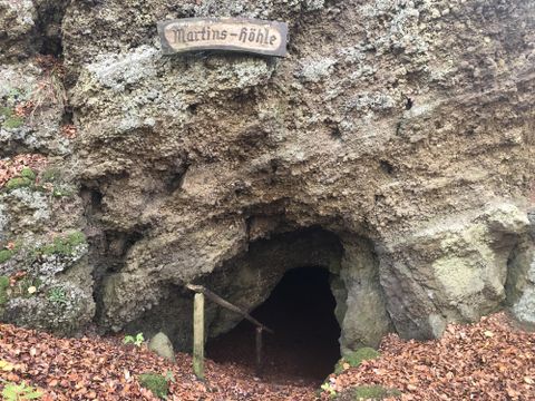 Entrance to the Martins Cave in Hohenfels-Essingen, surrounded by foliage and rocks. A wooden sign displaying the inscription 'Martins Cave' is visible.