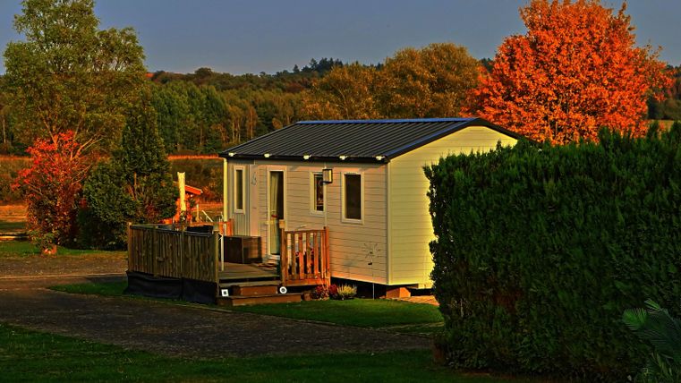 Ein modernes, weißes Ferienhaus mit einer Holzterrasse. Im Hintergrund leuchten die herbstlichen Bäume in orange und grün.