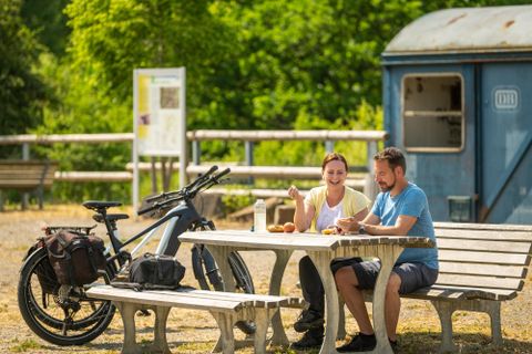 Twee personen zitten aan een picknicktafel op een rustplaats langs de Kyll-fietsroute. Een fiets staat ernaast, op de achtergrond is een oude DB-wagon te zien.