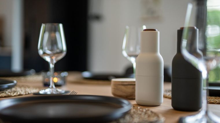 Ein elegant gedeckter Tisch mit dunklen Tellern und Gläsern. Auf dem Tisch stehen moderne Gewürzmühlen in Schwarz und Weiß sowie ein kleiner Behälter.