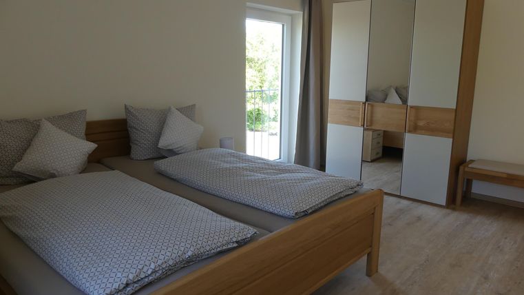 Ein helles Schlafzimmer mit zwei Betten, neutraler Dekoration und großen Fenstern. Ein Spiegelschrank und eine Holzbank ergänzen das gemütliche Ambiente.