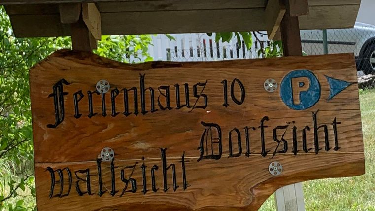 Ein Holzschild mit der Aufschrift „Ferienhaus 10 Waldblick“ und einem Parkplatzsymbol. Es ist von Bäumen umgeben und steht in einem Garten.