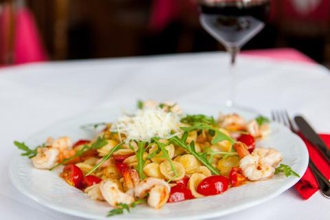 A plate with pasta, shrimp, arugula, and cherry tomatoes, garnished with grated cheese, on a white table with a glass of red wine.