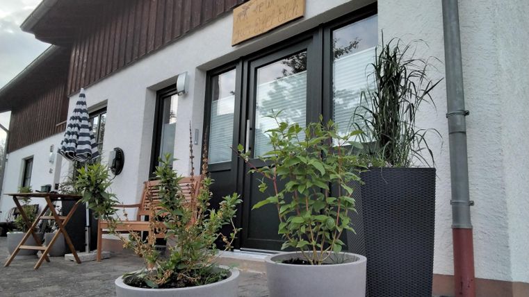 Ein modernes Gebäude mit großen Fenstern und einer einladenden Eingangstür. Vor dem Haus stehen einige Pflanzen in Töpfen und es gibt einen kleinen Tisch mit Stühlen.