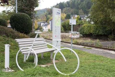 A creative bench in the shape of a bicycle stands at a bus stop. In the background, gentle hills and trees can be seen.