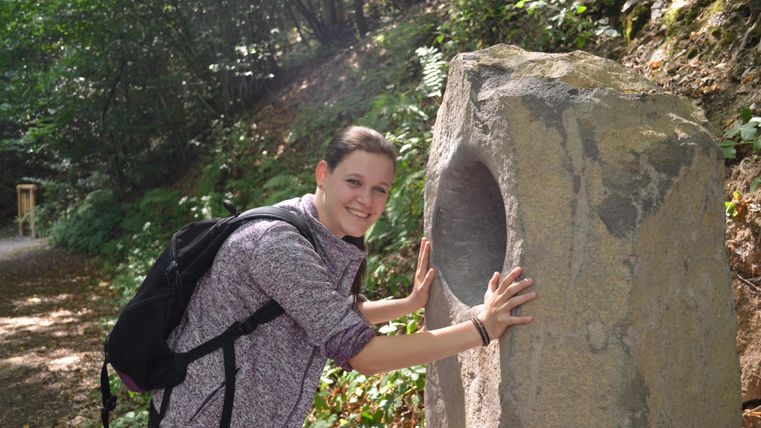 Eine Person mit einem Rucksack steht neben einem großen Stein mit einem Loch. Im Hintergrund sind Bäume und ein Wanderweg zu sehen.