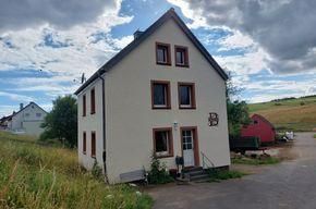 Ein modernes, zweigeschossiges Haus mit weißen Wänden und roten Fensterrahmen. Im Hintergrund sind grüne Wiesen und ein bewölkter Himmel zu sehen.