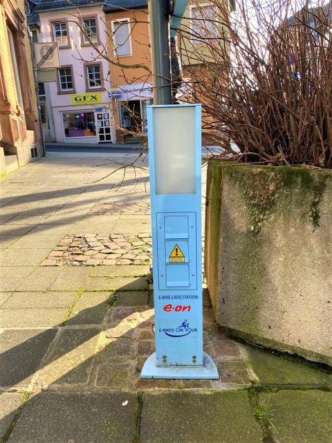 A blue charging station for electric vehicles stands on a sidewalk. In the background, several buildings and a small row of shops are visible.