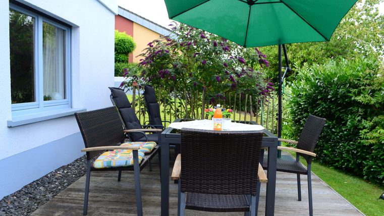 A cozy terrace with a table and chairs. A green umbrella provides shade and the garden is lushly overgrown.