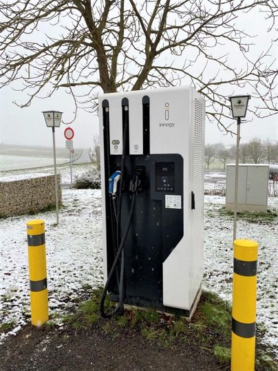 A modern charging station for electric vehicles stands in the snow. In the background, trees and a road are visible.