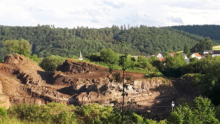 Ein abgebautes Gelände mit Erde und Steinen im Vordergrund. Im Hintergrund sind Bäume und Hügel zu sehen.