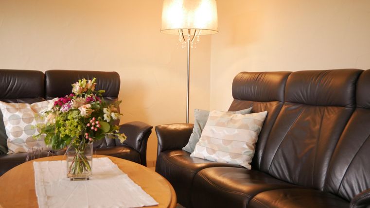 A cozy living room with two black leather sofas and a round wooden table. On the table, there is a vase with colorful flowers and a table lamp provides warm light.
