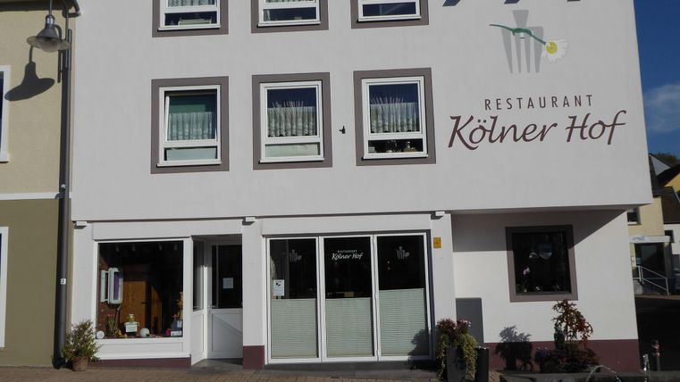 Ein Restaurant namens "Kölner Hof" mit einer hellen Fassade und großen Fenstern. Die Umgebung ist gepflastert und es gibt einige Pflanzen vor dem Eingang.