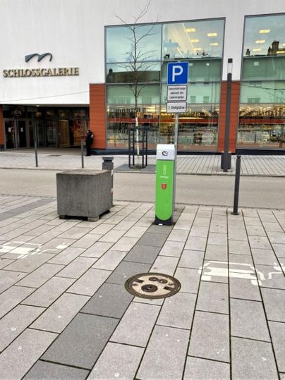 A parking lot with a modern parking meter near a shopping center. In the background, large windows and a sign with the name "Schildsgalerie" can be seen.