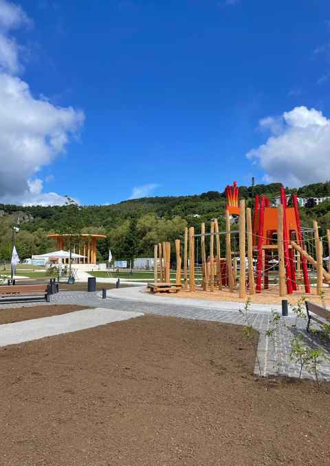 A new playground made of wood and with red elements is located on a paved area. In the background, trees and a wooded hill can be seen.