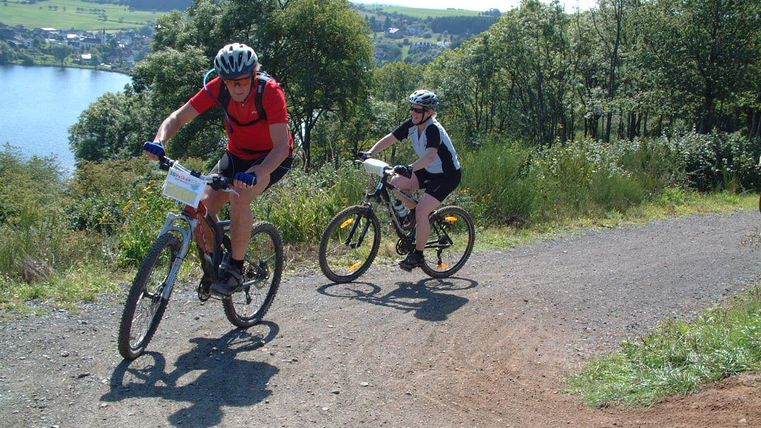 Two mountain bikers are riding up a steep path. In the background, a beautiful lake and a green landscape can be seen.