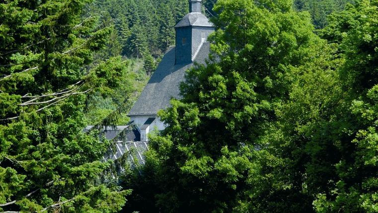 A monastery building with a tower, surrounded by dense green trees.