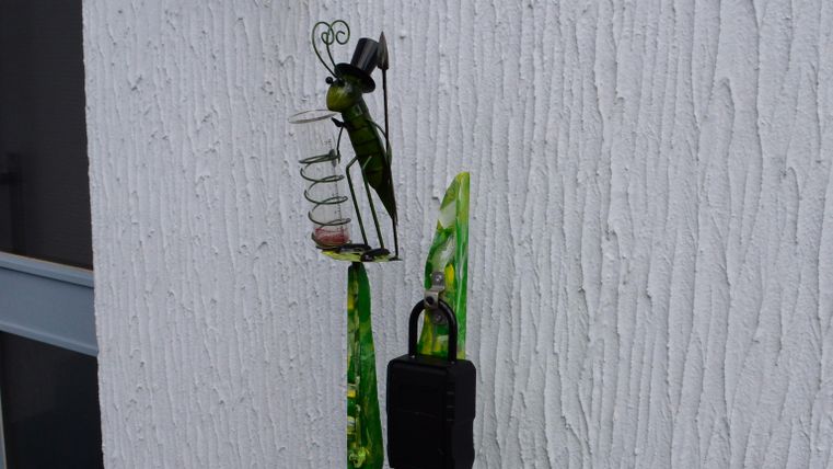 A decorative wall with a green, artistic column and a bird on a feeding station. Above that, a wall lamp is mounted.