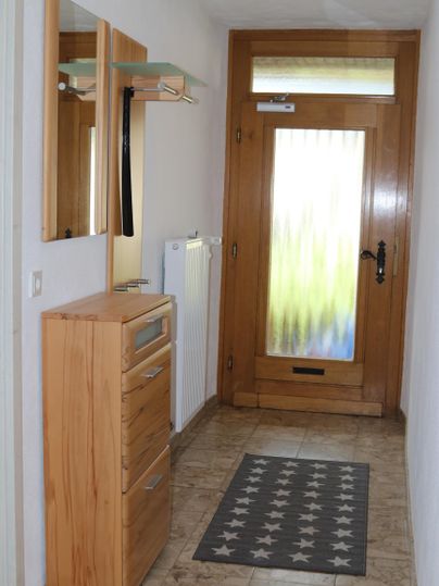 Ein heller Flur mit einer Holzkommode und einem Spiegel. Die Eingangstür hat eine große Glasfläche und die Wände sind in neutralen Farben gehalten.