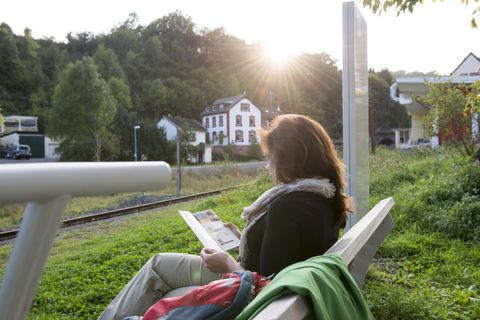 A woman is sitting on a bench and reading a magazine. In the background, the sun can be seen rising behind trees and a building.